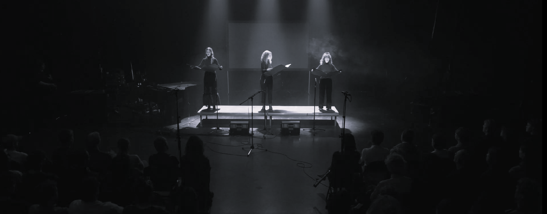 Performance still, three performers reading from lit scripts on stage